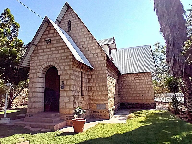 A historic stone building featuring a distinctive arched entrance, pitched corrugated metal roof, and established garden surroundings.