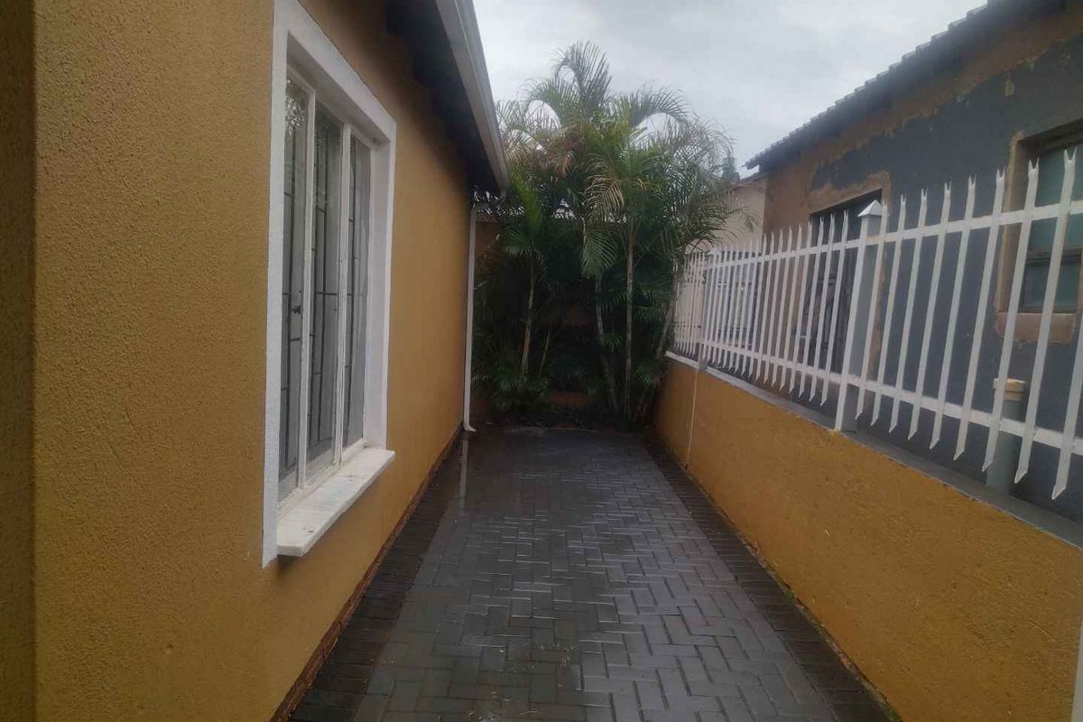 A narrow side passage featuring paved flooring, a white security fence, and exterior wall access to the property.