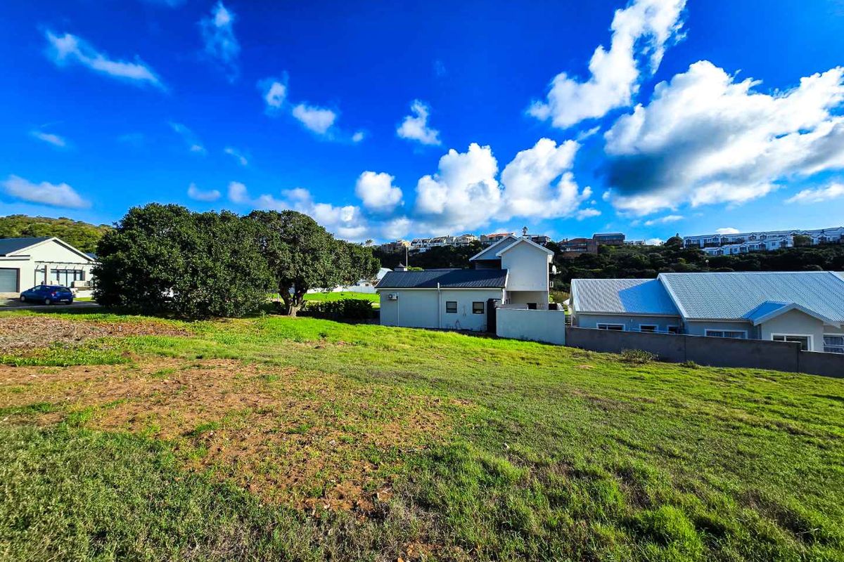A view of a vacant residential plot featuring grassy terrain, mature trees, and surrounding established residential properties under a clear blue sky.