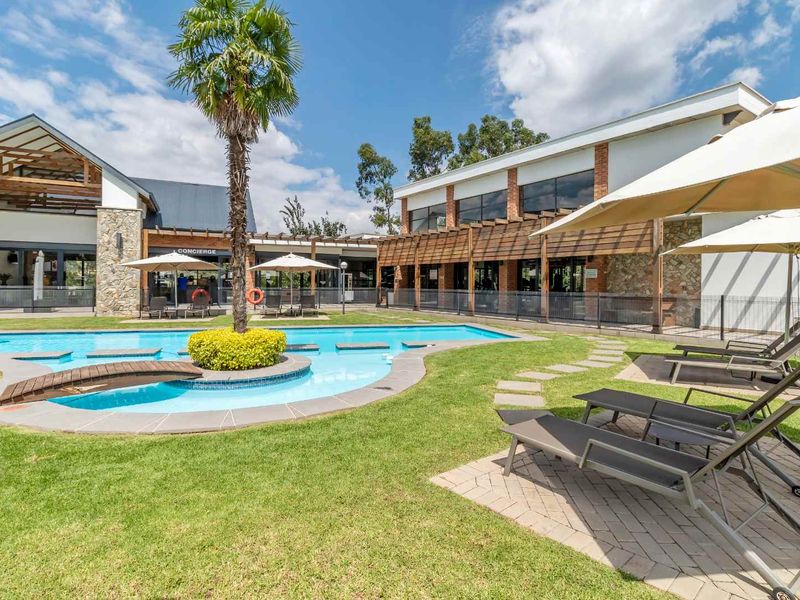 A well-maintained communal pool area featuring a landscaped garden, stone-clad building facades, timber pergolas, and paved lounging spaces.
