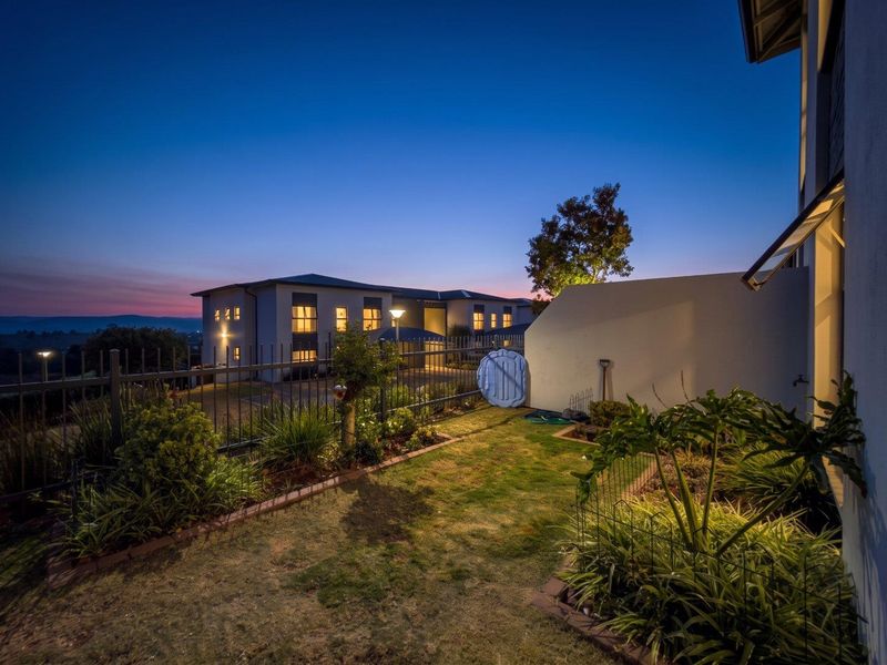 A well-maintained rear garden area featuring landscaped beds, metal perimeter fencing, and exterior lighting, set against a twilight backdrop of the apartment complex.