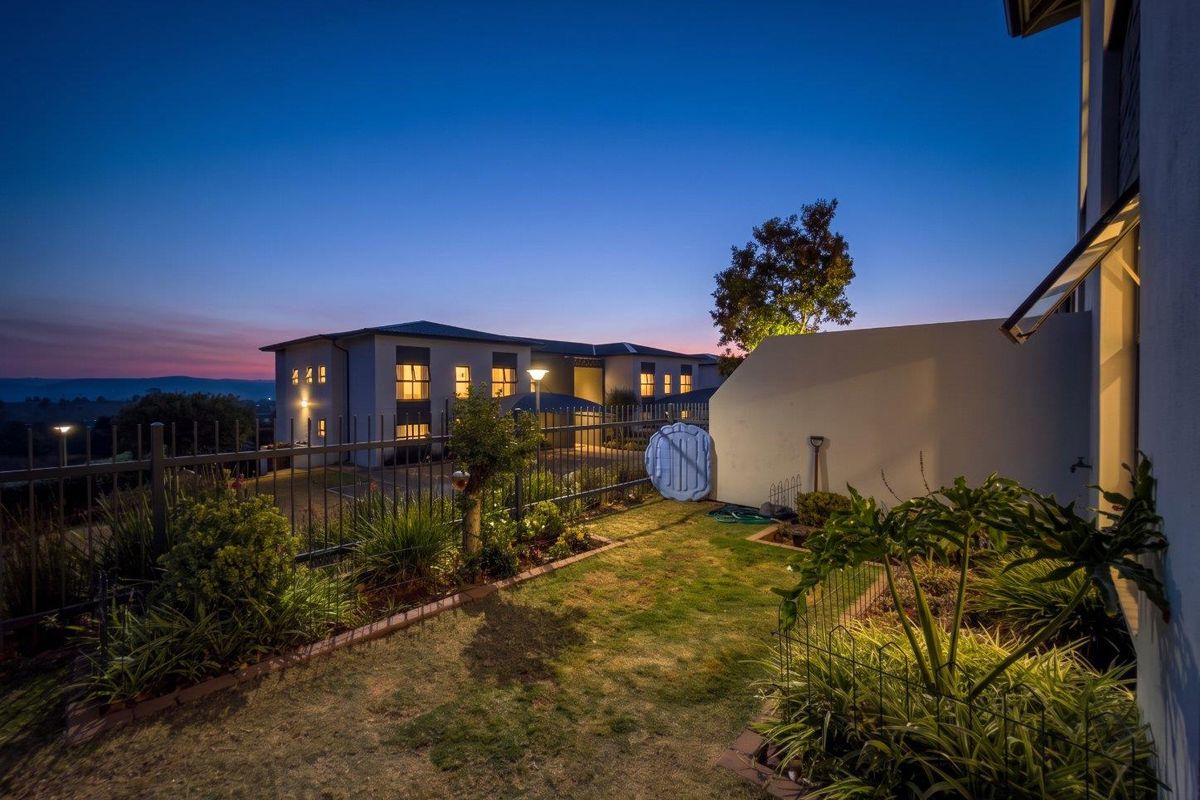 A well-maintained rear garden area featuring landscaped beds, metal perimeter fencing, and exterior lighting, set against a twilight backdrop of the apartment complex.