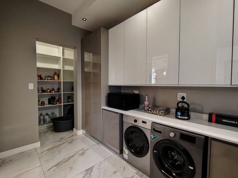 Modern scullery featuring sleek high-gloss cabinetry, stone countertops, integrated laundry appliances, and a spacious walk-in pantry with ample shelving.