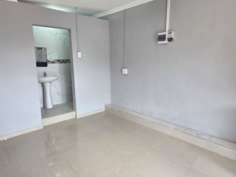 An empty open-plan living area featuring tiled floors, exposed electrical conduit, and an adjoining small wash area with a pedestal basin.