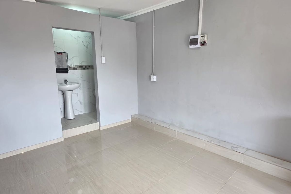 An empty open-plan living area featuring tiled floors, exposed electrical conduit, and an adjoining small wash area with a pedestal basin.