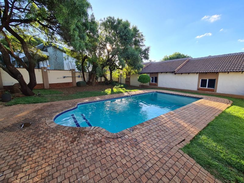 A well-maintained outdoor pool area featuring a paved deck, surrounding lawn, and perimeter wall, set against a residential building with a tiled roof.