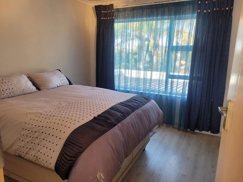 A functional bedroom featuring light-toned laminate flooring and a large window with curtains, providing natural light and a view of the exterior.