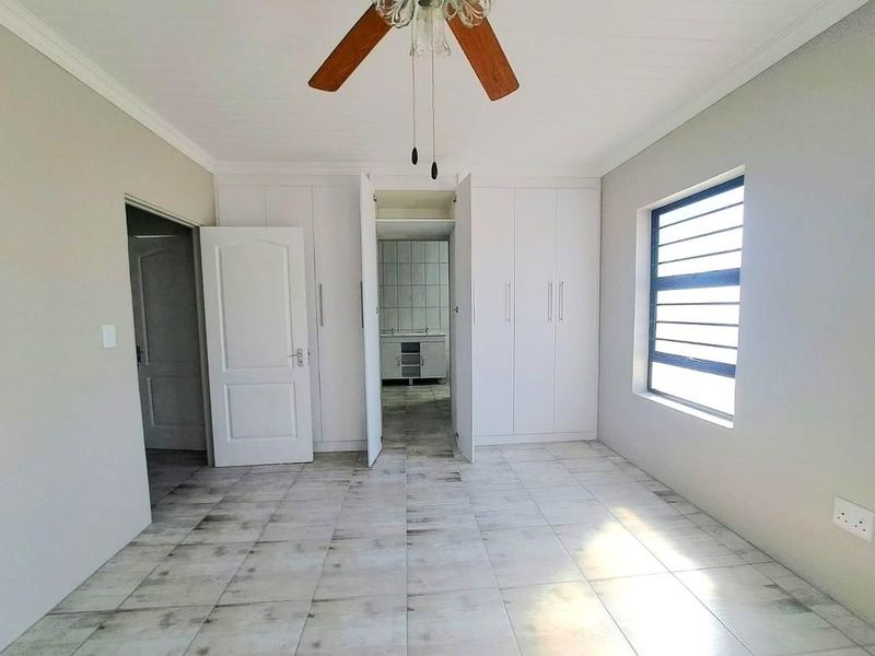 A spacious, empty bedroom featuring built-in cupboards, tiled flooring, a ceiling fan, and a view through to an adjacent tiled utility area.