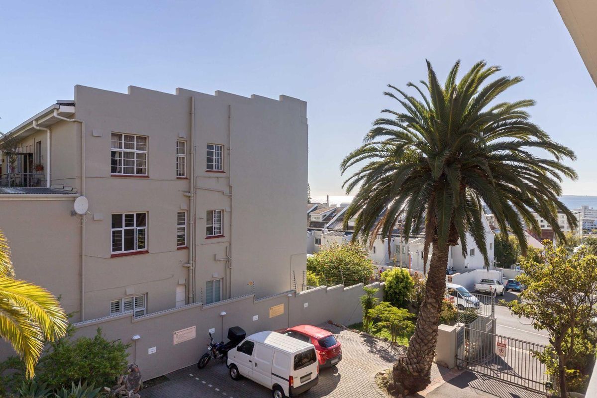 View of the apartment building exterior, featuring a paved driveway, perimeter wall with security fencing, and mature palm tree in the rear grounds.