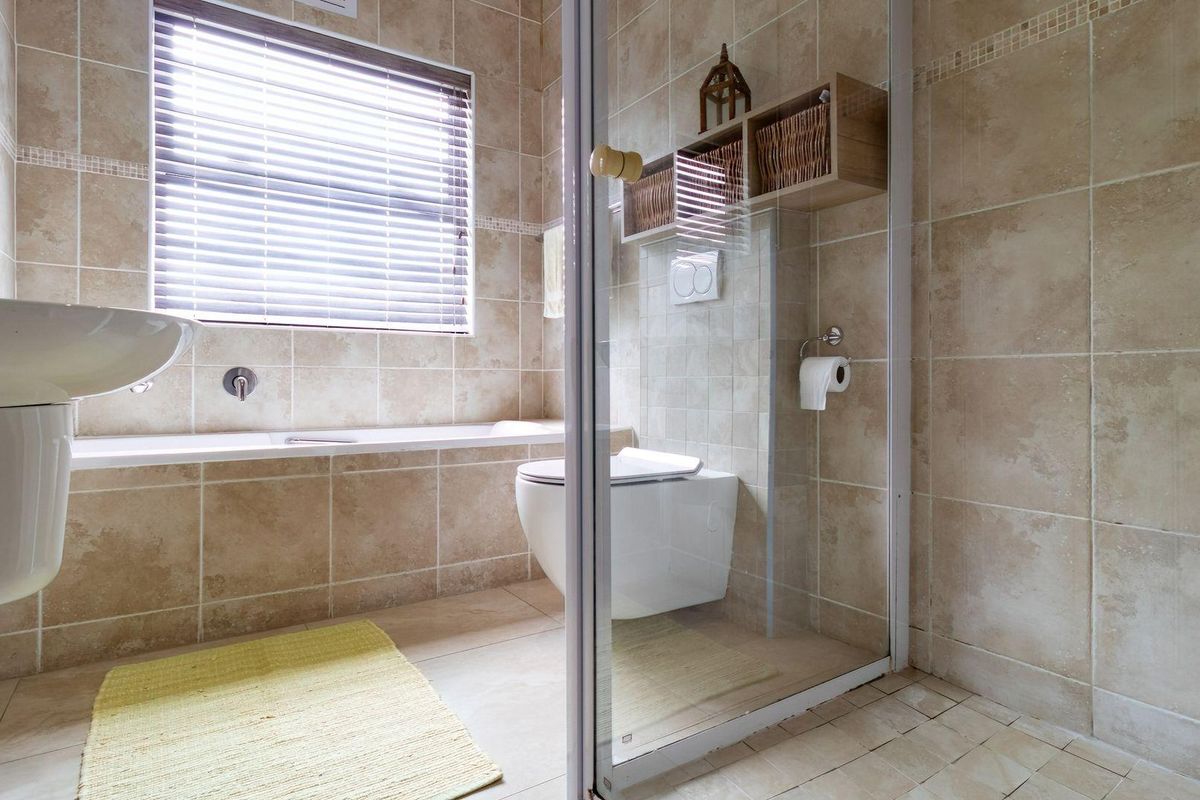 A modern bathroom featuring floor-to-ceiling beige tiling, a wall-mounted toilet, a glass-enclosed shower, and a built-in bathtub under a window.