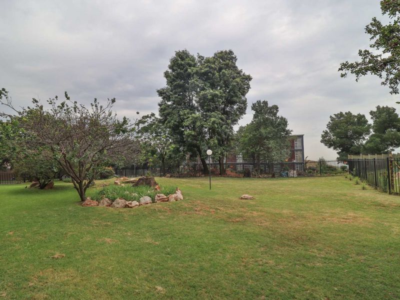 A spacious communal garden area featuring a well-maintained lawn, mature trees, and secure perimeter fencing.