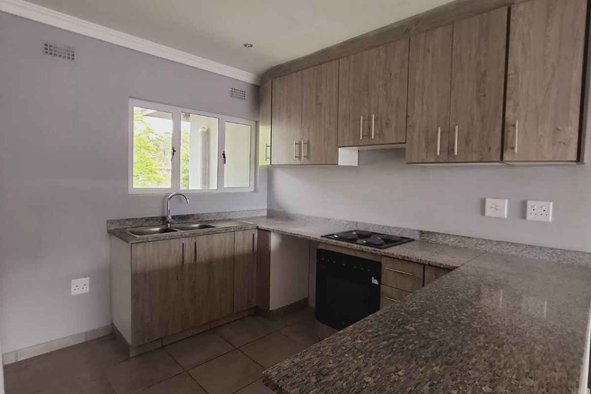 A clean, modern kitchen featuring wood-finish cabinetry, granite countertops, built-in oven and hob, and tiled flooring in a well-lit, open-plan layout.