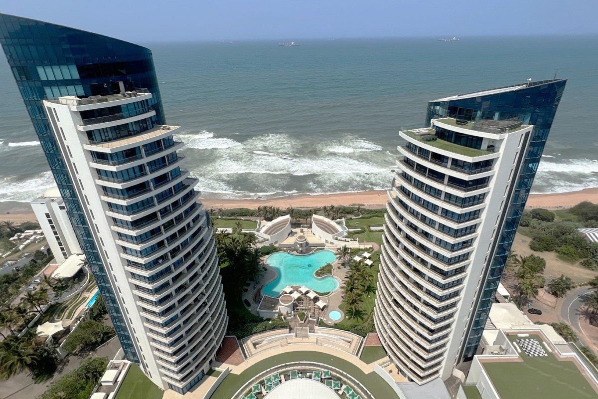 Aerial view of a modern luxury apartment complex featuring twin high-rise towers, a central swimming pool, and direct oceanfront access.