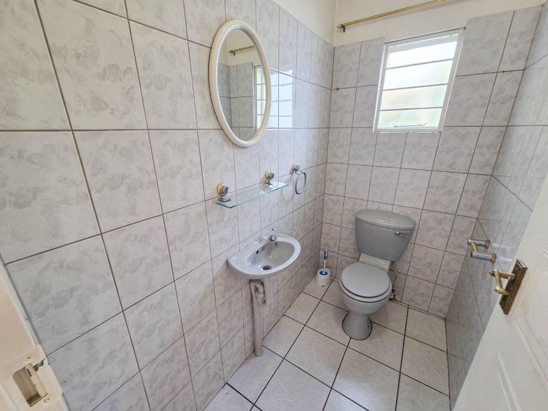 A compact, fully tiled bathroom featuring a wall-mounted basin, toilet, and louvered window, presenting a functional and dated aesthetic.