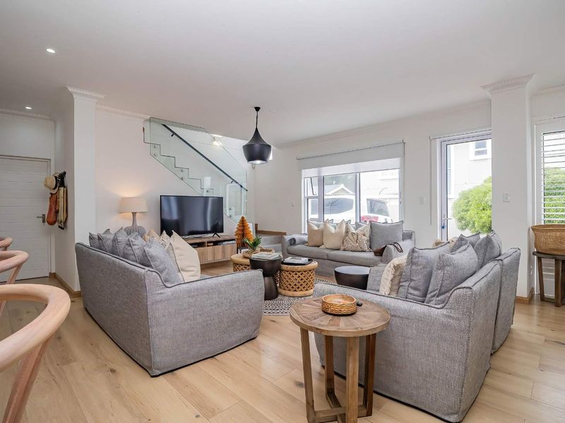 A bright and modern living room featuring light hardwood flooring, a contemporary glass balustrade staircase, and ample natural light through large windows and doors.