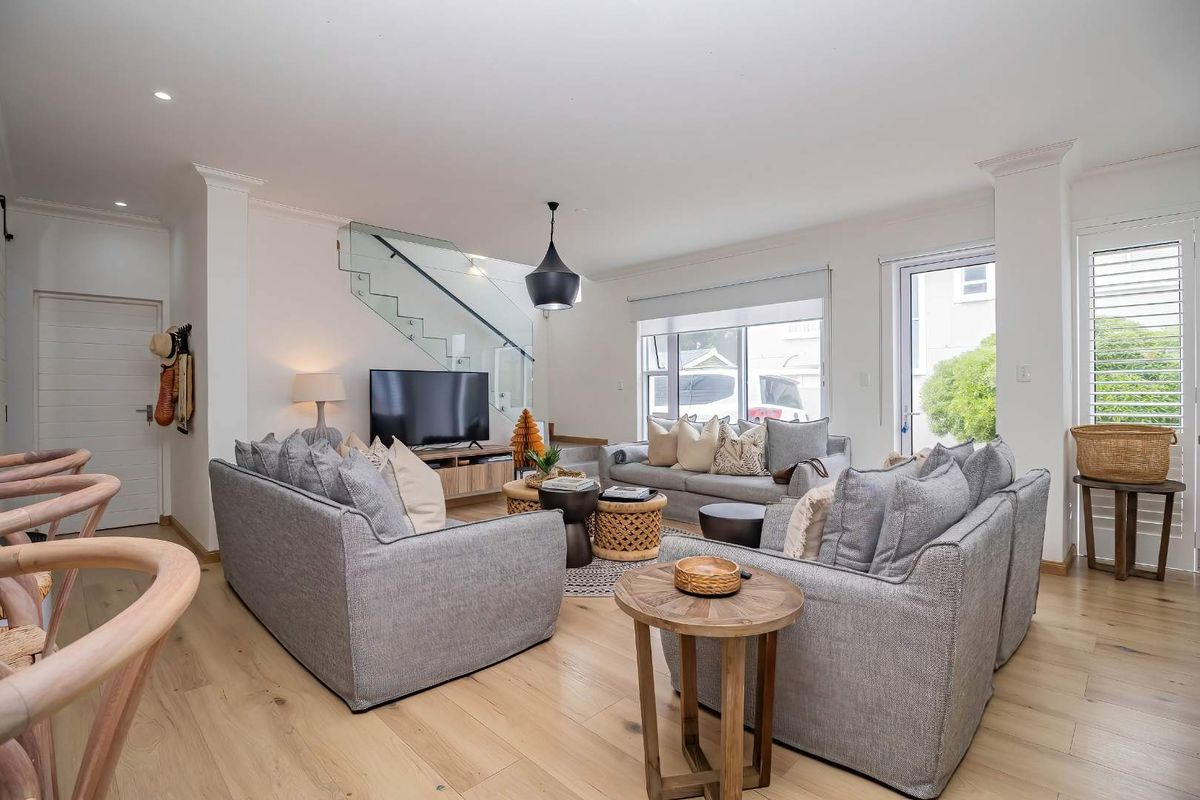 A bright and modern living room featuring light hardwood flooring, a contemporary glass balustrade staircase, and ample natural light through large windows and doors.