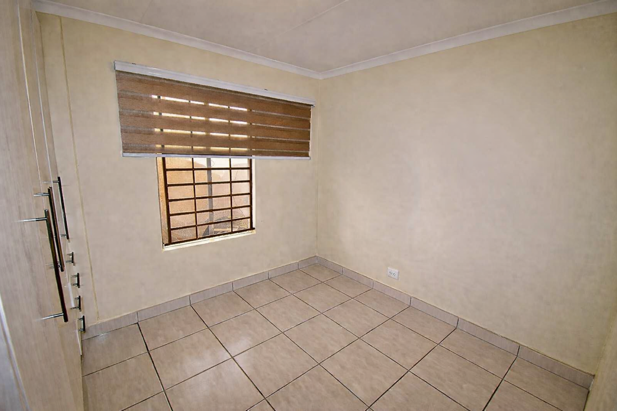 A compact, empty bedroom featuring neutral floor tiles, built-in cupboards, and a window fitted with a security gate and roller blind.
