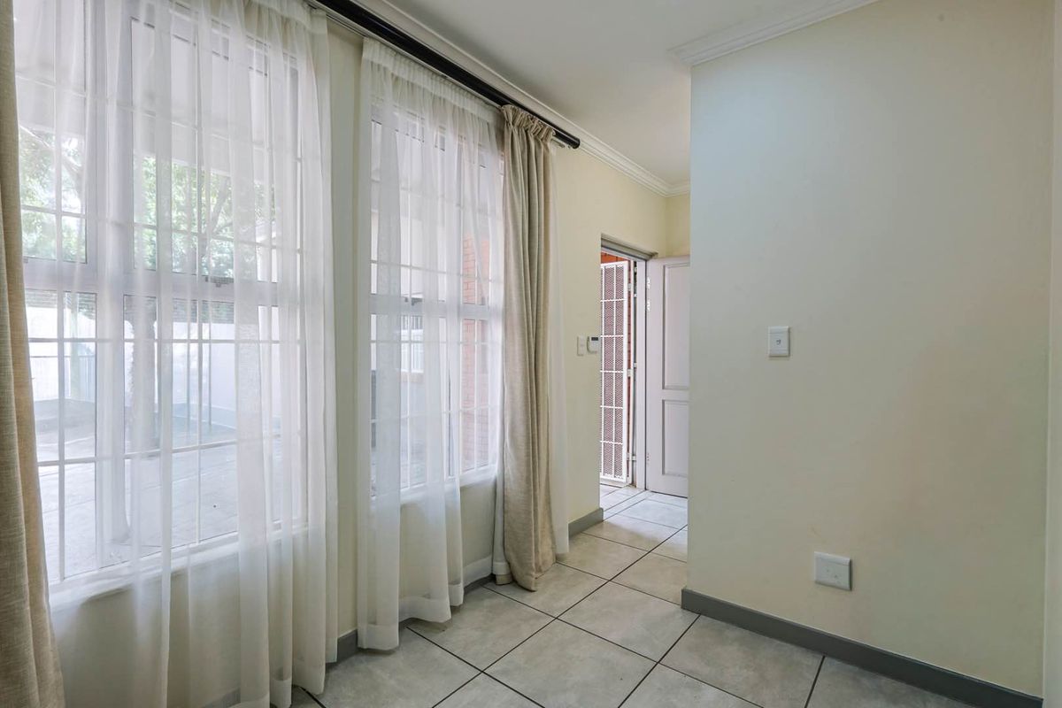 A bright, tiled interior space featuring large windows with sheer curtains, leading to an adjacent room through a doorway.