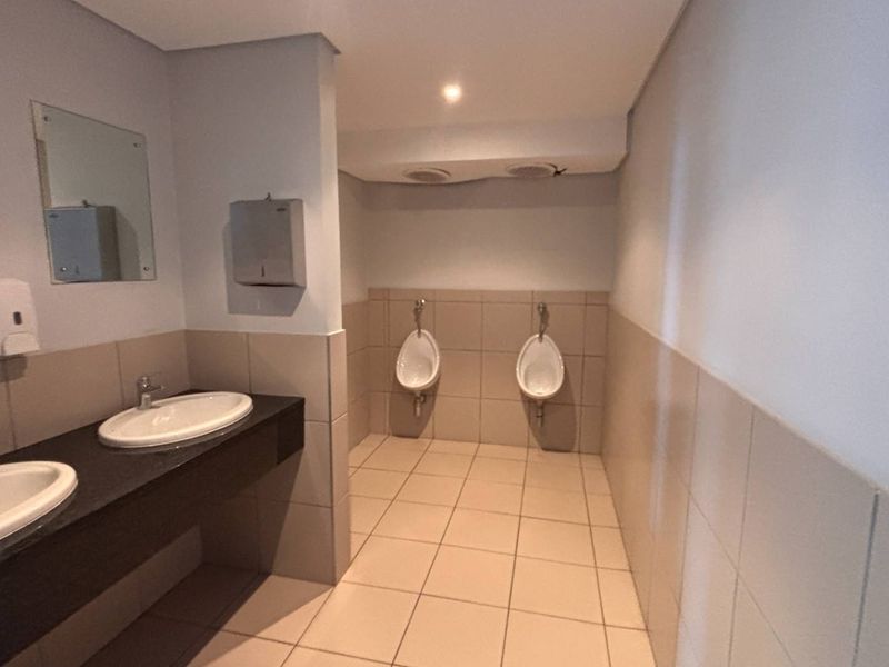 A clean, functional office restroom featuring dual vanity basins, wall-mounted urinals, and neutral tiled finishes throughout.