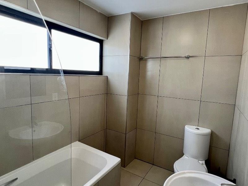 A clean, modern bathroom featuring neutral wall and floor tiles, a bathtub with a glass screen, a pedestal basin, and a toilet.