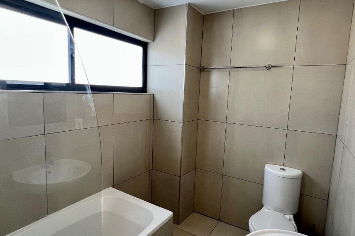 A clean, modern bathroom featuring neutral wall and floor tiles, a bathtub with a glass screen, a pedestal basin, and a toilet.