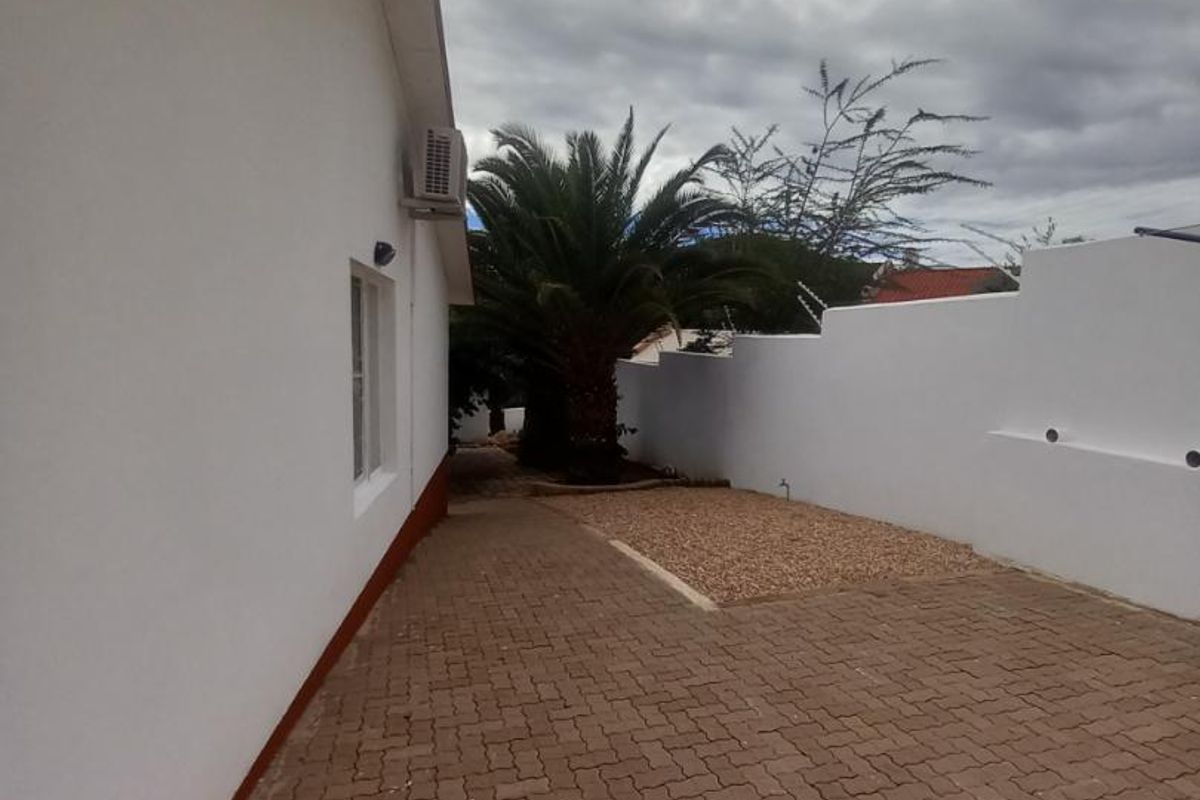 A side exterior view of the property featuring paved driveway, white boundary wall, and established palm tree.