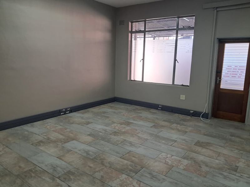 A clean, empty office space featuring modern wood-look tiled flooring, neutral wall paint, and functional aluminium window frames.