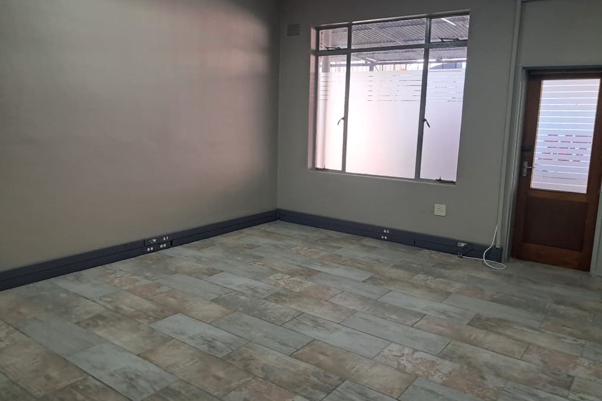 A clean, empty office space featuring modern wood-look tiled flooring, neutral wall paint, and functional aluminium window frames.