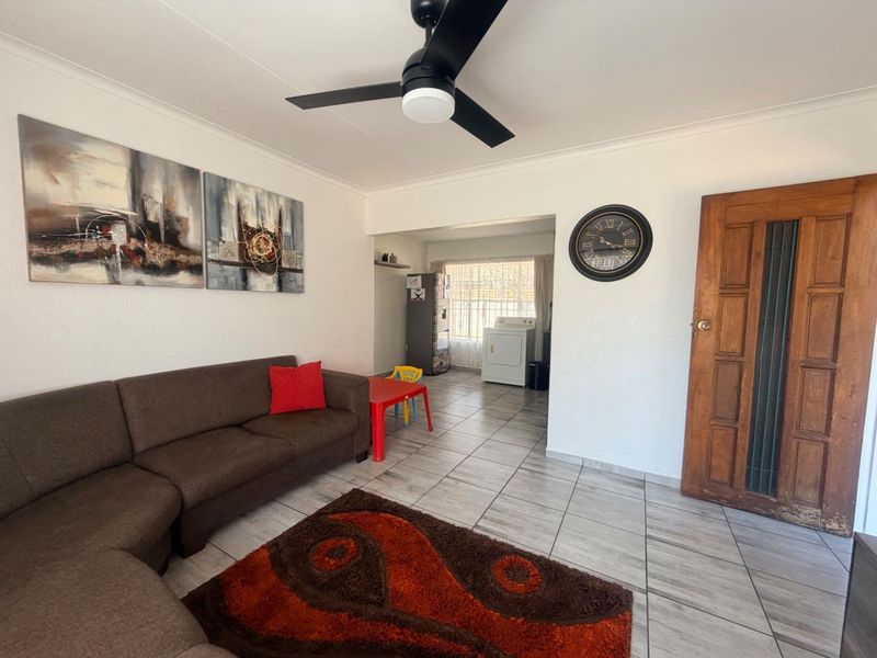 Open plan living area featuring tiled flooring, a ceiling fan, and a solid wooden door leading to adjacent utility space.