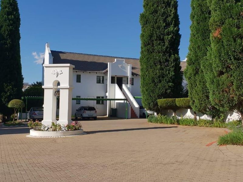 A well-maintained apartment complex entrance featuring a paved driveway, mature cypress trees, and covered parking bays.