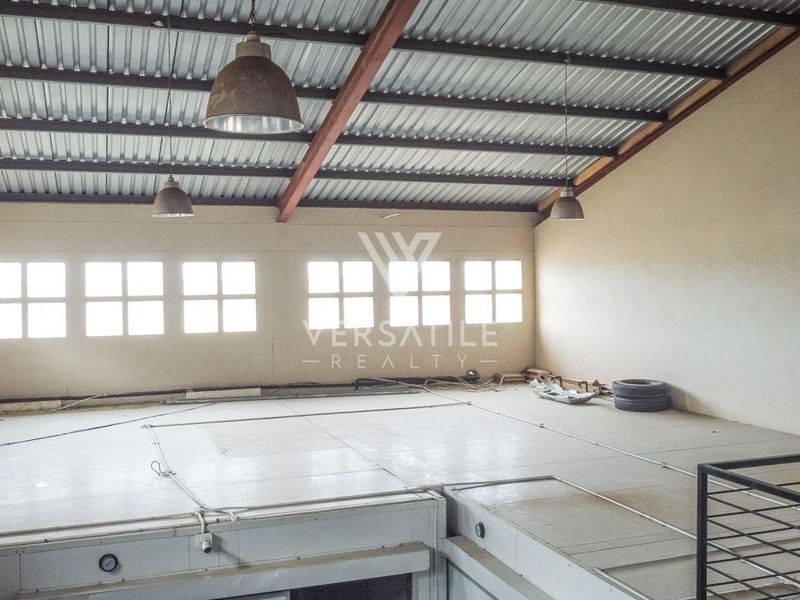 Spacious industrial mezzanine area featuring exposed steel roof trusses, industrial pendant lighting, and large grid windows, suitable for storage or office conversion.