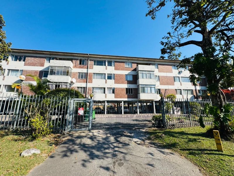 Front view of a multi-story apartment building featuring a brick facade, secure gated entrance, and covered ground-level parking.