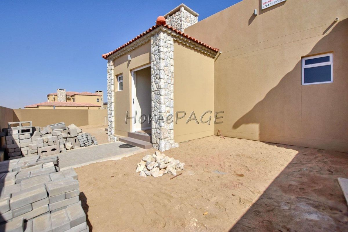 Exterior view of a house entrance featuring stone-clad pillars, a tiled roof, and an unfinished sandy yard with construction materials present.