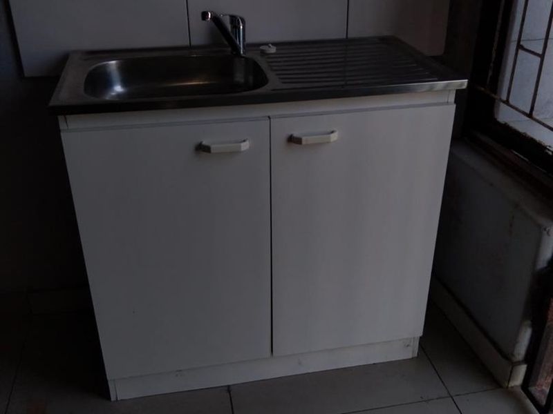 A basic kitchen sink unit with white cabinetry and tiled splashback, set against an unfinished wall in a tiled room.