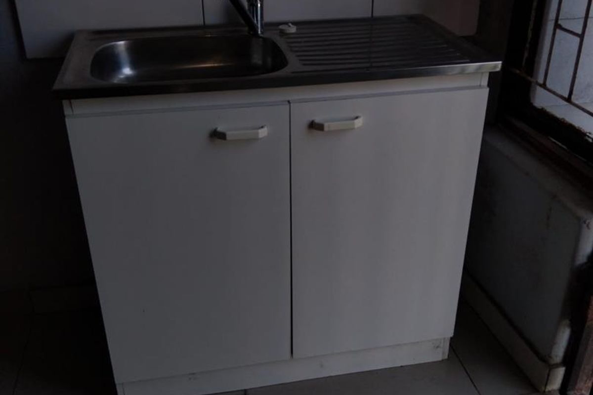 A basic kitchen sink unit with white cabinetry and tiled splashback, set against an unfinished wall in a tiled room.