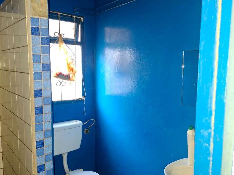 A small, basic bathroom featuring a toilet, wall-mounted basin, and a tiled shower partition, requiring significant cosmetic updates and floor finishing.