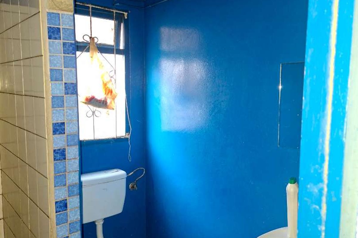 A small, basic bathroom featuring a toilet, wall-mounted basin, and a tiled shower partition, requiring significant cosmetic updates and floor finishing.