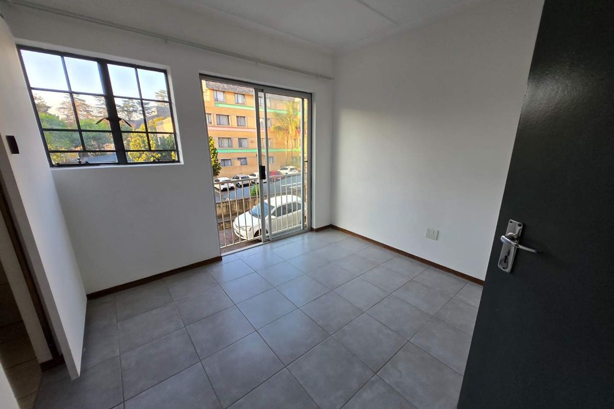 A bright, empty bedroom featuring neutral tiled flooring, a sliding glass door leading to a balcony, and large windows providing natural light.