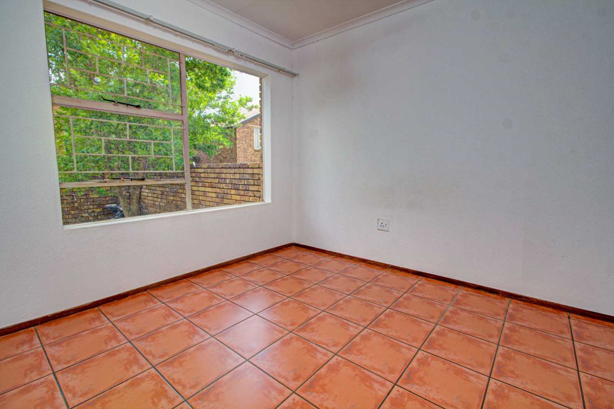An empty bedroom featuring terracotta floor tiles, a large window with security bars, and plain white walls.
