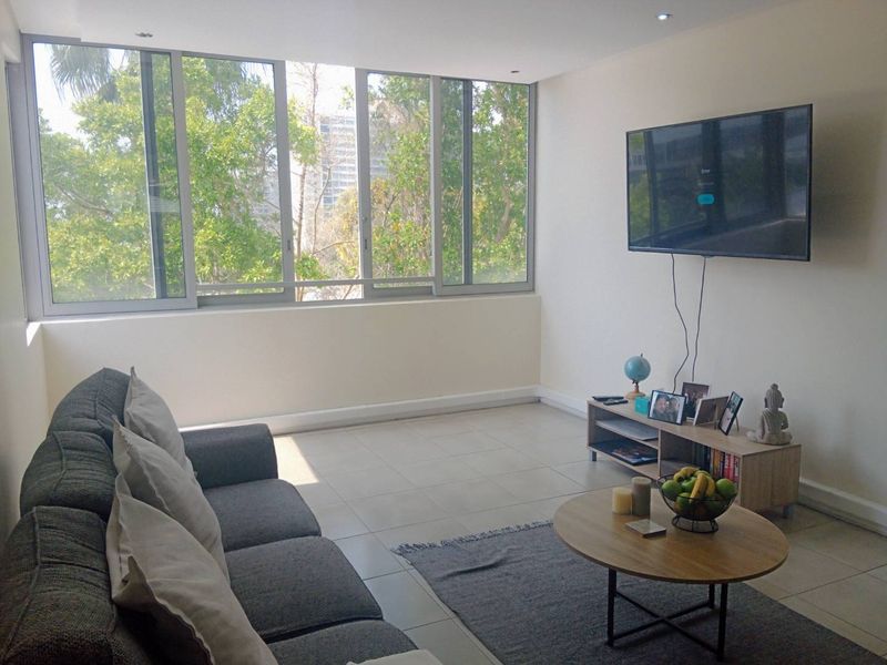 A bright living room featuring large sliding windows with natural views, neutral tiled flooring, and recessed ceiling lighting.
