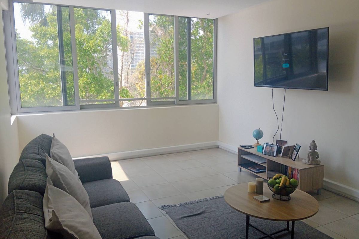 A bright living room featuring large sliding windows with natural views, neutral tiled flooring, and recessed ceiling lighting.
