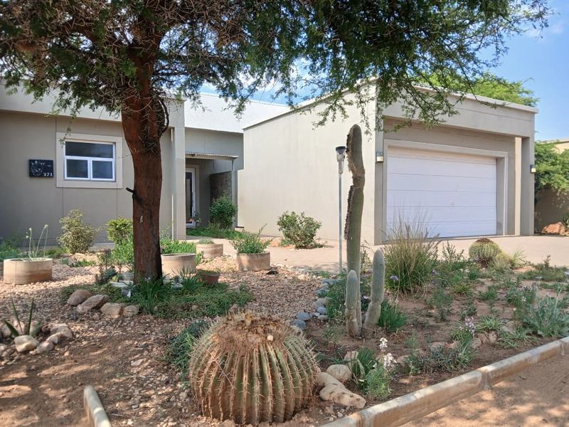 Front elevation of a modern house featuring a double garage, paved driveway, and a well-maintained xeriscape garden with mature trees and cacti.