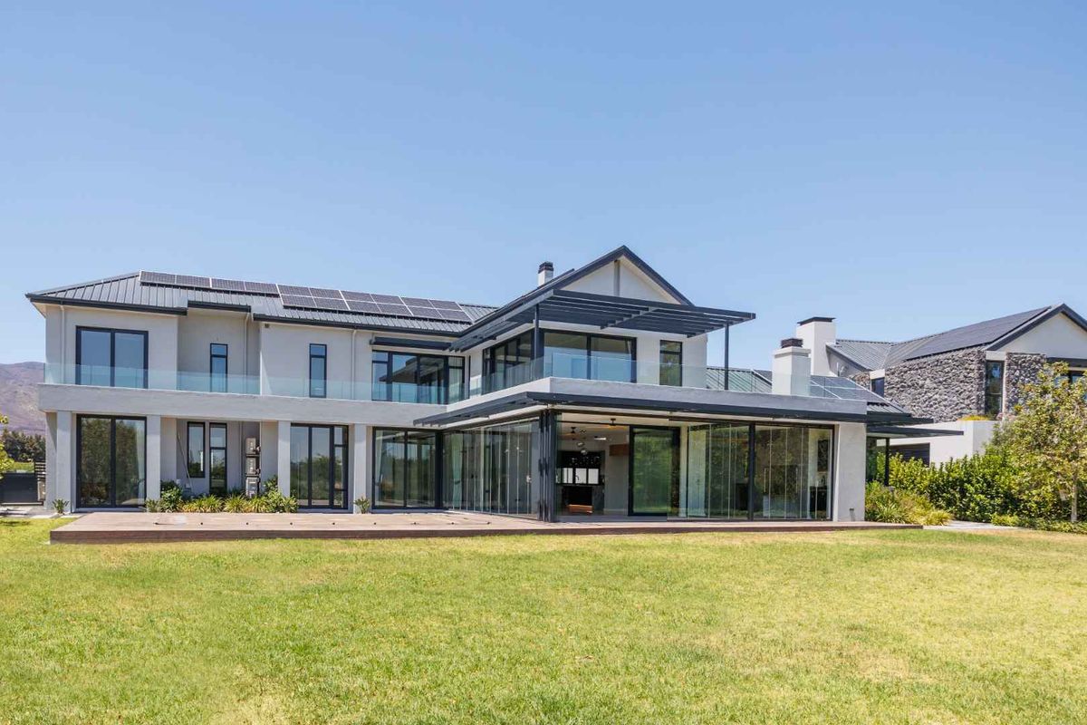 Rear view of a modern two-story residence featuring extensive glass sliding doors, timber decking, solar panels, and a spacious landscaped lawn.