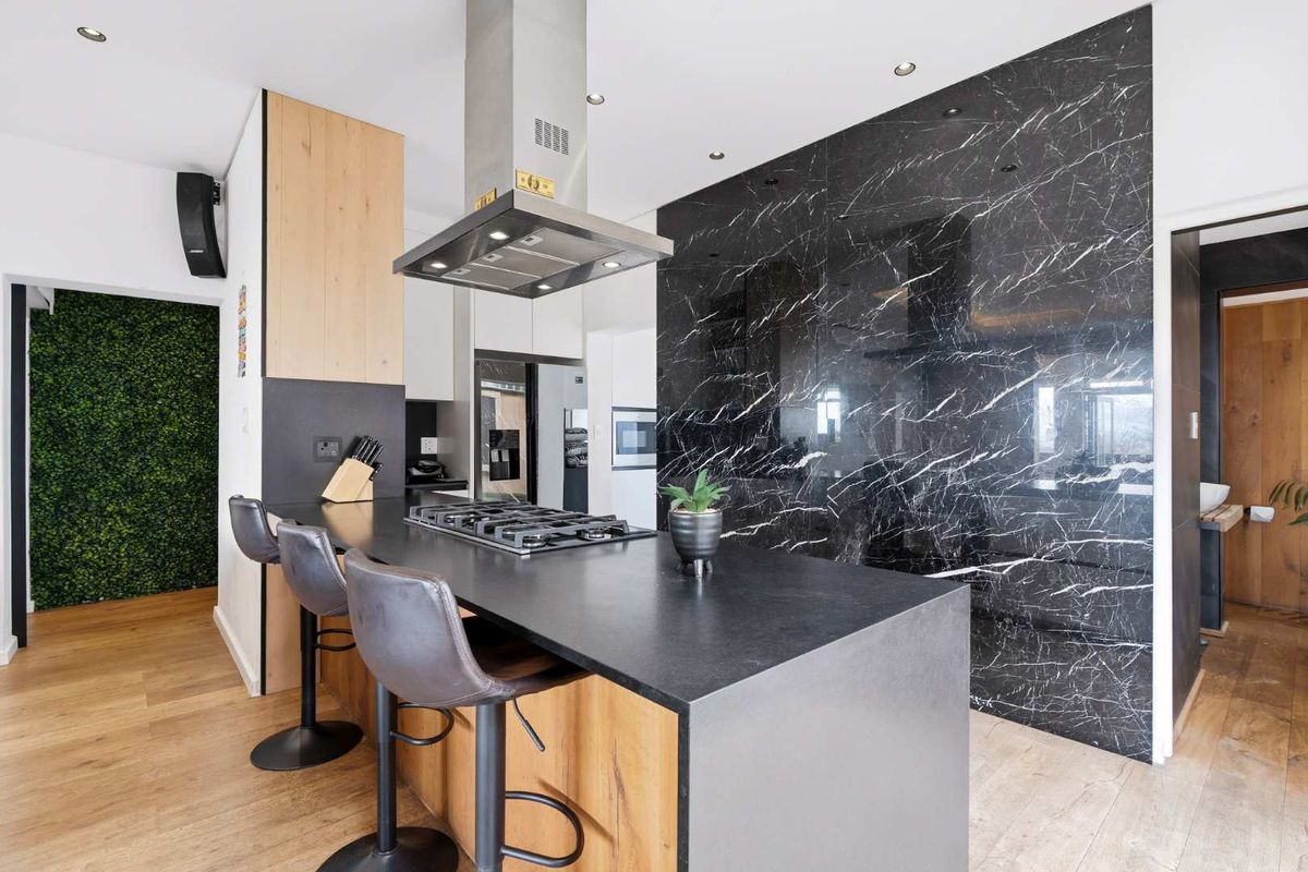 Modern kitchen featuring a central island with gas cooktop, stainless steel extractor fan, and striking black marble-look splashback with warm timber cabinetry accents.