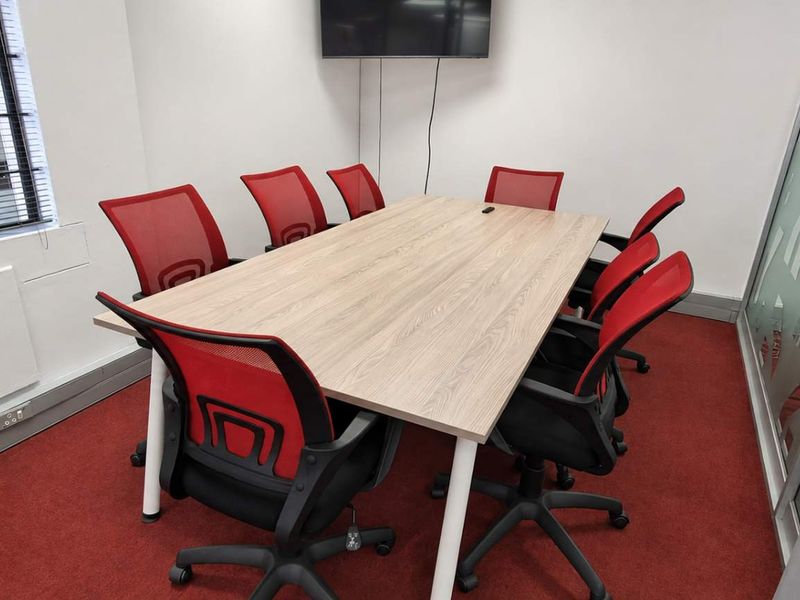 A functional office meeting room featuring a central conference table, red ergonomic chairs, a wall-mounted screen, and a glass partition wall.