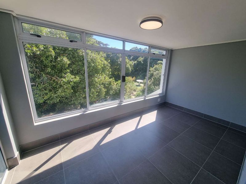 A bright, empty enclosed balcony or sunroom featuring large windows with garden views and modern tiled flooring.