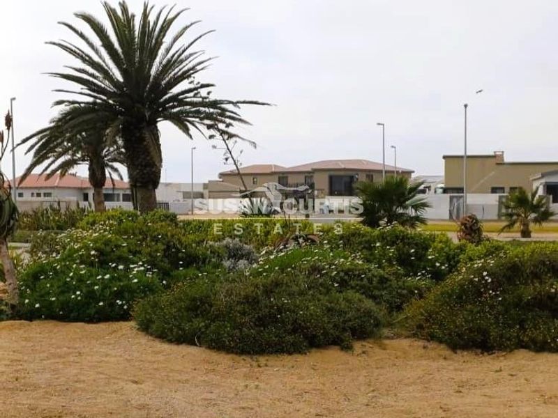 A street-level view of the property exterior featuring landscaped garden beds, mature palm trees, and surrounding residential infrastructure.