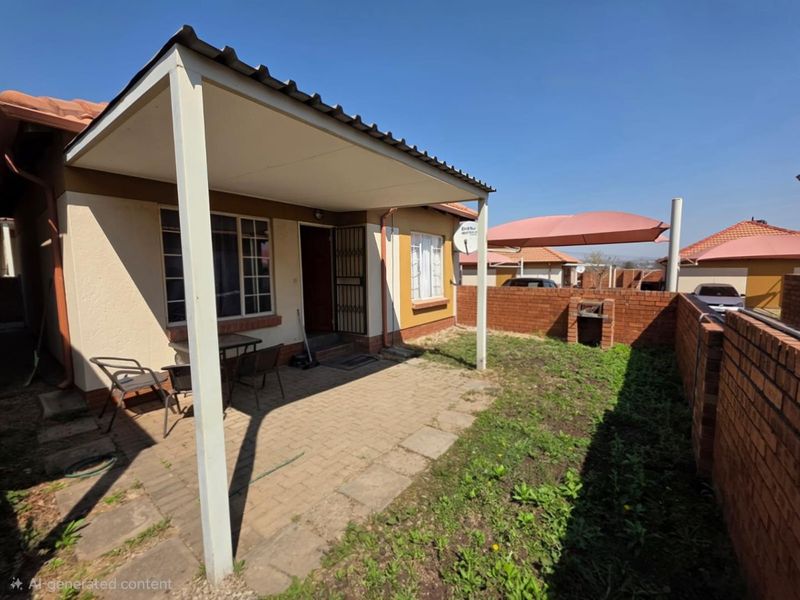 Rear exterior view of a townhouse featuring a covered patio, paved area, and a built-in brick braai within a private garden space.
