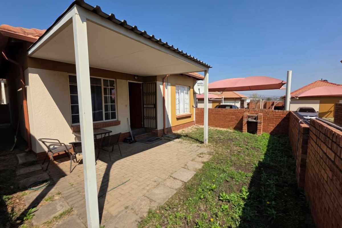 Rear exterior view of a townhouse featuring a covered patio, paved area, and a built-in brick braai within a private garden space.