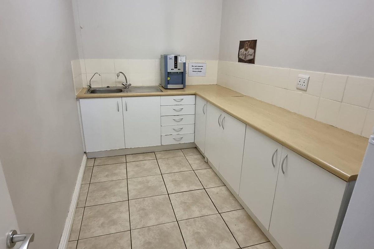 A functional office kitchenette featuring white built-in cabinetry, a laminate countertop, stainless steel sink, and tiled flooring.